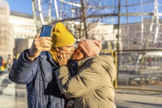 Happy senior couple kissing and taking a selfie with smartphone in a winter city during christmas