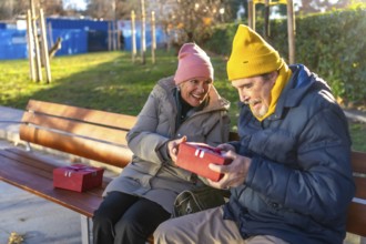 Elderly man receiving christmas gift from smiling wife while sitting on bench in city park