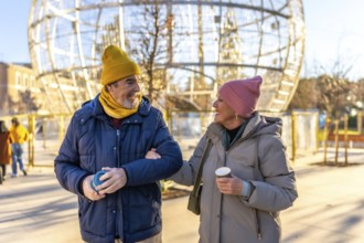 Smiling senior couple walking arm in arm in a city during winter, enjoying a warm beverage and the