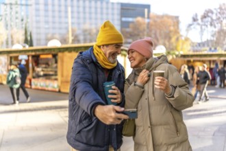 Smiling senior couple taking a selfie and drinking hot beverages at a christmas market in the city