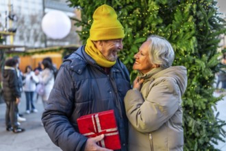 Elderly man surprising his wife with a christmas present at a christmas market during winter