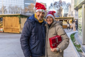 Smiling senior couple wearing santa hats holding christmas present at a christmas market in the