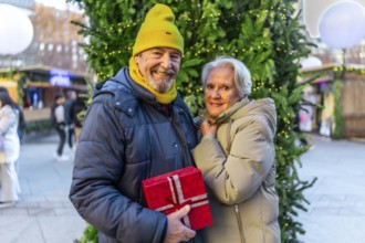 Smiling senior couple holding christmas gift, enjoying christmas market in the city during winter