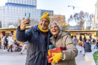Cheerful senior couple taking a selfie with smartphone at a christmas market, holding gifts and