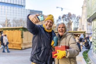 Elderly couple taking a selfie with smartphone at a christmas market, holding gifts and hot drinks,