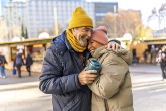 Elderly couple hugging and smiling at an outdoor christmas market, enjoying a warm beverage on a
