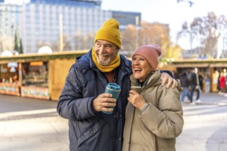 Smiling senior couple enjoying hot drinks at an outdoor christmas market, embracing the festive
