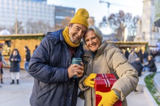 Smiling senior couple holding christmas gift and hot beverage at outdoor christmas market, enjoying