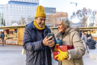 Happy retired couple looking at smartphone and holding christmas gifts and reusable coffee cup at a