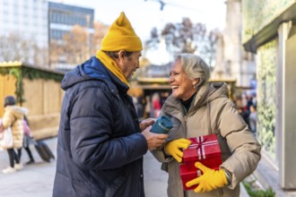 Smiling elderly couple exchanging christmas presents and enjoying hot beverages at a winter market,