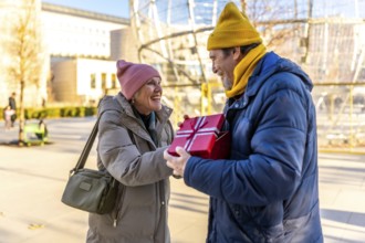 Elderly man joyfully giving a christmas present to a smiling woman in a sunny winter city park,