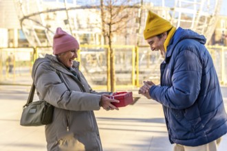 Cheerful senior woman giving a christmas present to her excited husband in a city during wintertime