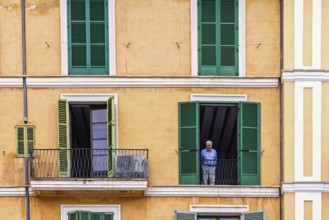 Senior man standing and looking out of an open window in an apartment house, Palma de Mallorca,
