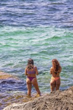 Young women at a rocky beach standing in the water's edge by the sea, Mallorca, Spain