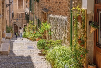 Alone woman walking in an idyllic cobblestone alley in an old village with green plants and flowers