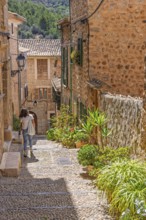 Woman walking in an idyllic cobblestone alley in an old village with green plants and flowers in
