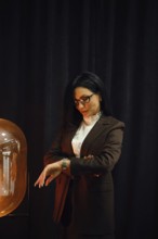 A woman dressed in a fashionable brown suit stands near a glowing lamp. She checks her watch with a