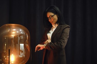 A confident woman in a tailored brown suit stands beside a stylish lamp. She checks her watch