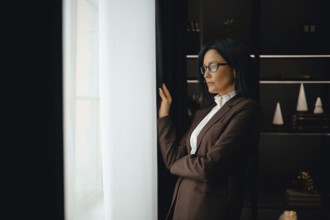 A professional woman dressed in a brown suit stands thoughtfully by a window, gazing outside