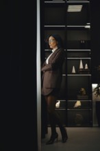 A confident woman in brown jacket, mini skirt and glasses stands with her arms crossed in an office