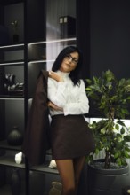 A professional woman stands confidently in office, wearing a white blouse and a brown skirt. She