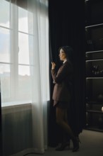 A woman dressed in a tailored suit stands by a large window, pulling back sheer curtains. She