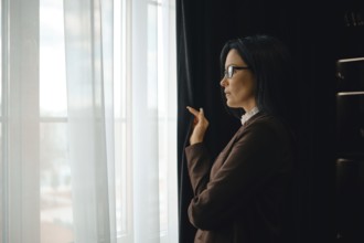 A businesswoman stands by a window in a modern office, gazing thoughtfully outside. She wears