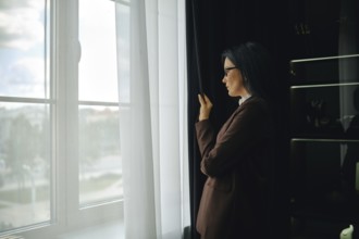Woman in brown jacket gazes out window while reflecting on the day in a modern setting