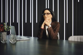 A thoughtful woman with glasses sits at a modern office desk. She has long dark hair and is dressed
