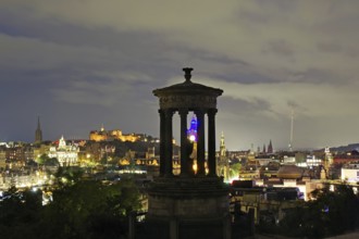 Night scene of Edinburgh with illuminated buildings and a historical monument in the foreground,