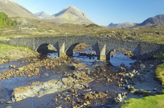 Stone bridge over a shallow river in the middle of a mountain landscape under a clear sky,