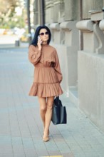 A fashionable woman strolls down the city sidewalk wearing a brown dress and sunglasses, carrying a
