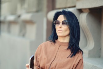 A woman wearing stylish sunglasses stands next to a historic building, casually holding her