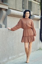A relaxed woman stands by a textured stone wall, wearing a stylish brown dress with a flowing skirt