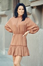 A woman stands with her arms on waist on a city sidewalk, wearing a chic, loose-fitting dress. Her