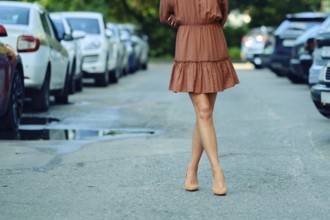 Unrecognizable woman is walking down a city street, wearing a brown dress and high heels. The