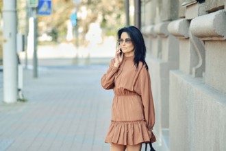 A fashionable woman dressed in a brown dress engages in a phone conversation while standing on a
