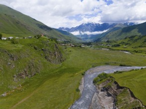 River flowing through green valley, surrounded by mountains and clouds, near a small village,