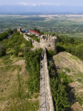 Historic castle wall with towers and a path, surrounded by lush vegetation and a wide view of the