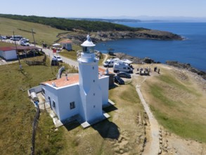 A lighthouse on the coast with mobile homes nearby, surrounded by meadow and rocks, the sea horizon