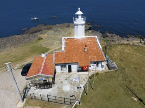 A lighthouse on a rocky coast with blue sky and sea, next to it a building with a terracotta roof,