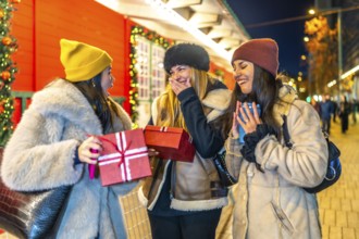 Three cheerful women exchanging christmas gifts and enjoying hot beverages at a vibrant night