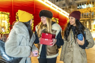 Three cheerful young women holding christmas gifts and enjoying a winter night out in a decorated