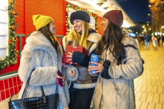 Three cheerful women holding christmas gifts and hot drinks, enjoying christmas shopping at a