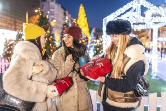 Three joyful women exchanging christmas gifts in a beautifully decorated city square, surrounded by