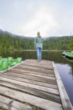 Woman standing alone on a wooden jetty in front of a quiet lake surrounded by forest, Mummelsee,