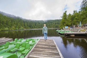 Woman on a jetty at the lake, surrounded by forest and boats under a cloudy sky, Mummelsee,