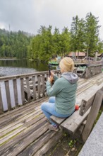 Woman taking a photo of the lake while sitting on a wooden bench surrounded by forest, Mummelsee,