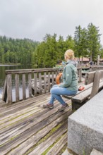 A woman sits relaxed on a wooden bench overlooking the tranquil lake and forest, Mummelsee,