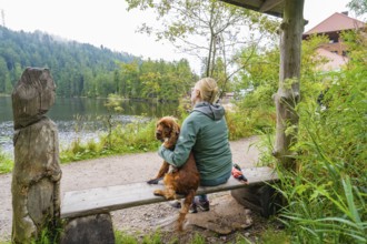 Woman sitting with dog on a bench by the lake in the forest and enjoying the tranquillity of